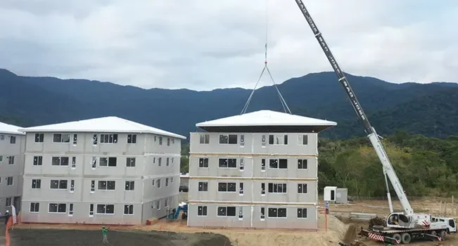 Um grande guindaste eleva a cobertura de um prédio de quatro andares no canteiro de obras do Conjunto Habitacional São Sebastião. Dois edifícios semelhantes se completam ao fundo, com uma paisagem montanhosa além. Trabalhadores e equipamentos são visíveis no terreno