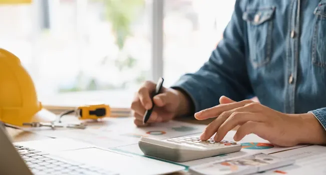 Homem sentado a mesa, com caneta em uma mão e calculadora na outra. Ele verifica quais análises econômicas serão tendência na construção civil em 2025.